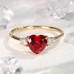 May include: A gold ring featuring a heart-shaped, deep red gemstone flanked by two smaller, round, clear gemstones. The ring's band is a thin, polished gold. The gemstones are set against a soft, white background.