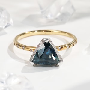Op de afbeelding: Een gouden ring met een driehoekige, donkerblauwe edelsteen. De edelsteen is omgeven door kleine diamanten en gezet in een wit metaal. De gouden band heeft kleine, gelijkmatig verdeelde diamanten.