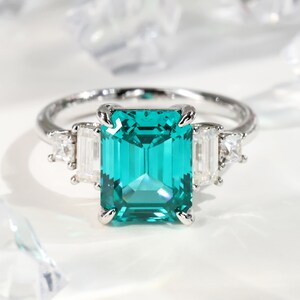 May include: A silver ring featuring a large, emerald-cut, teal gemstone as the centerpiece. Two rectangular and two triangular white gemstones flank the central stone. The ring is set against a white background.