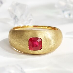 May include: A gold ring with a brushed finish, featuring a square, vibrant red gemstone at the center. The ring has a wide band and a smooth, rounded shape, showcasing a classic and elegant design.