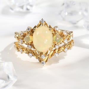 Può includere: Un set di anelli color oro con una pietra centrale di opale ovale e piccoli accenti di opale. L'anello è adornato da piccoli diamanti scintillanti e presenta un design a forma di foglia. Il set include una fascia abbinata.