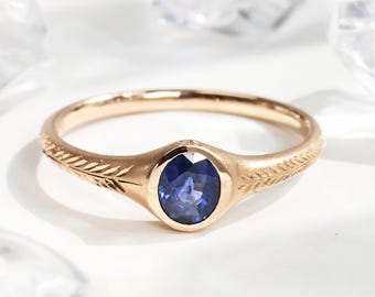 May include: A gold ring with an oval, deep blue gemstone at the centre. The band features a delicate leaf pattern on both sides of the gemstone. The ring is set against a white background, with crystal-like objects in the background.