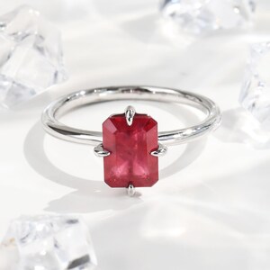 Puede incluir: Un anillo de plata con una gema rectangular de color rojo intenso. El anillo tiene una banda fina y pulida, y la gema está sujeta por cuatro pequeñas garras. La gema es el punto focal, reflejando la luz y mostrando un color vibrante.