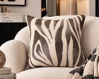 May include: A square throw pillow featuring a dark brown and cream zebra stripe pattern. The pillow is positioned on a cream-colored textured armchair, which is draped with a black and cream striped throw blanket with fringe detailing.