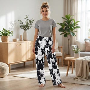 May include: Black and white pajama pants with a repeating pattern of cat faces. The cats are stylized with large eyes and simple features. The pants have a black waistband and a loose, comfortable fit. The pattern is evenly distributed across the fabric.