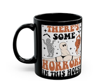 Op de afbeelding: Zwarte keramische mok met een Halloween-thema ontwerp. Het ontwerp toont een mummie, een spook en een pompoen, samen met de tekst "THERE'S SOME HORRORS IN THIS HOUSE" in oranje en zwart.