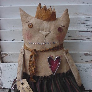 Könnte beinhalten: Handgemachte Katzendoll mit Krone, Schal und herzförmiger Verzierung. Die Katze hat Knopfaugen, eine genähte Nase und ein Etikett mit der Aufschrift "THE QUEEN". Der Rock der Puppe ist grün und braun gestreift.
