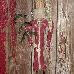 Op de afbeelding: Een folk art kerstmanpop met een lange rode jas en een witte baard. De pop houdt een kleine groene tak vast en draagt een geruite hoed. De pop is gemaakt van hout en stof, tegen een rustieke houten achtergrond.