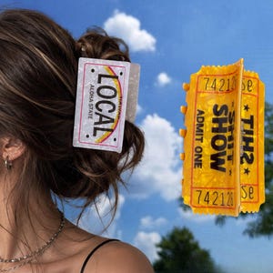 May include: A white hair claw clip featuring a rectangular design that mimics a Hawaii license plate with the text LOCAL ALOHA STATE, and a yellow hair claw clip shaped like an admission ticket with the text ADMIT ONE SHIT SHOW 742128.