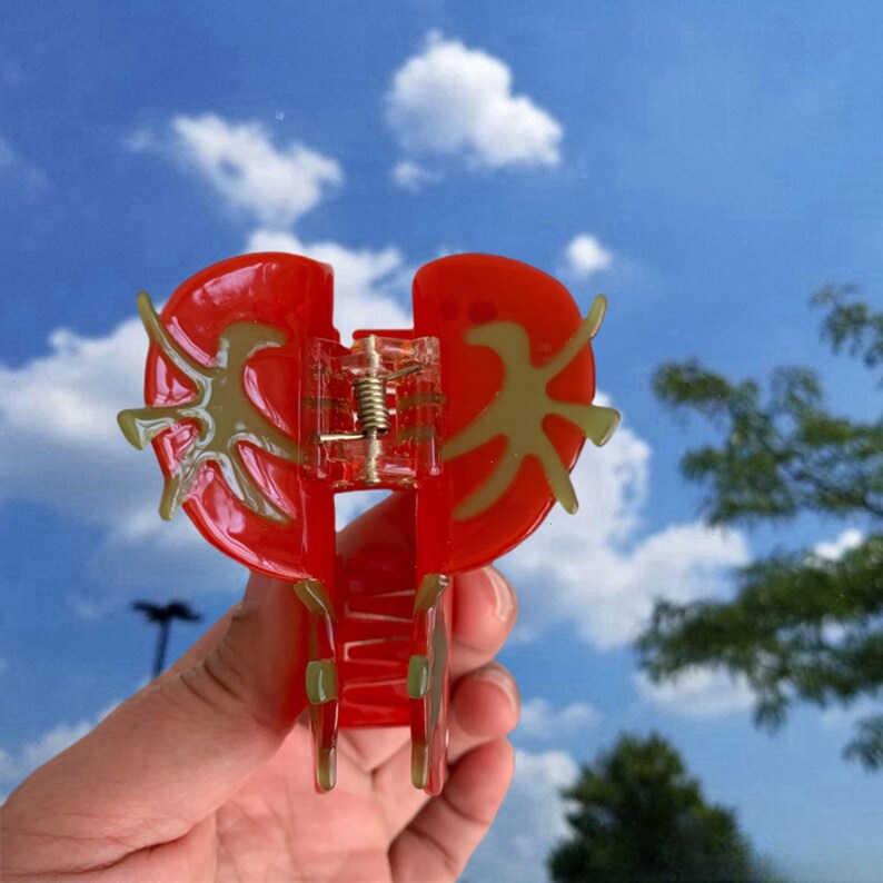 May include: A red heart-shaped hair clip with a gold design. The clip is open and ready to be used.