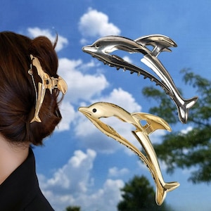 Puede incluir: Tres pinzas para el pelo de delf&iacute;n en oro y plata. Las pinzas tienen forma de delf&iacute;n con la cola enroscada. Las pinzas est&aacute;n sobre un fondo de cielo azul.
