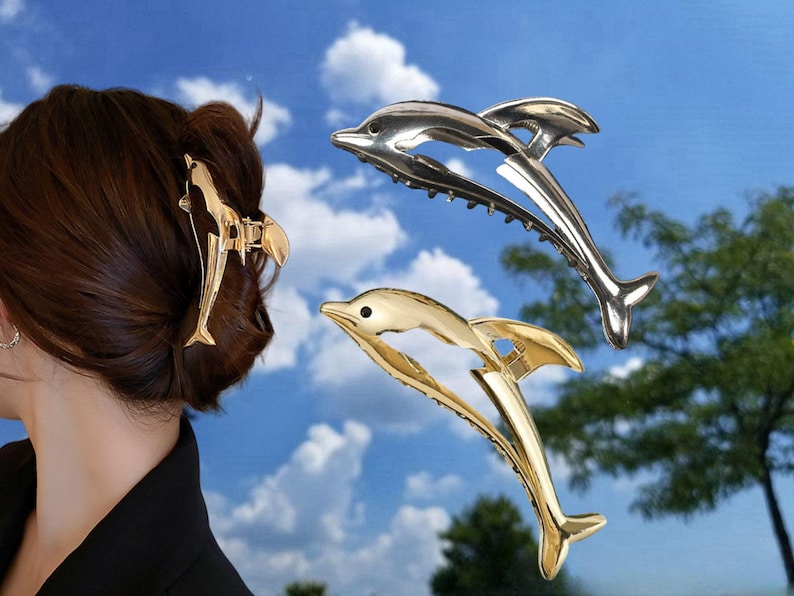 Puede incluir: Tres pinzas para el pelo de delf&iacute;n en oro y plata. Las pinzas tienen forma de delf&iacute;n con la cola enroscada. Las pinzas est&aacute;n sobre un fondo de cielo azul.