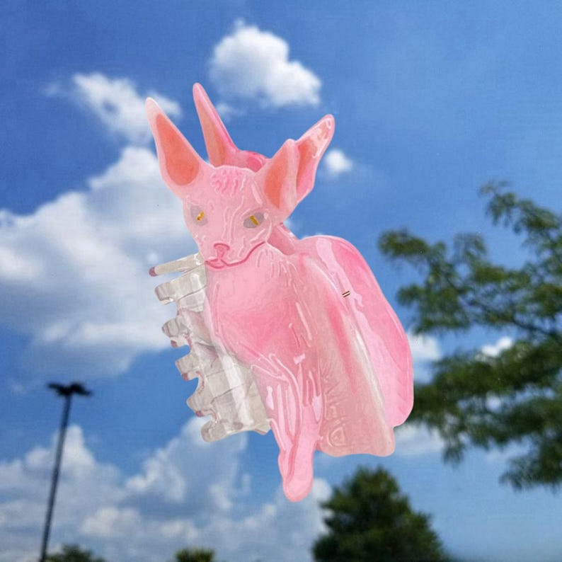 May include: A translucent pink hair claw clip shaped like a Sphynx cat with large pointed ears and yellow eyes. The accessory features a clear plastic spring mechanism with interlocking teeth designed to hold hair securely in place.