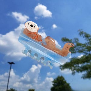 May include: A light blue hair claw clip featuring a brown sea otter design. The clip has a translucent blue base with a smooth finish and a series of teeth along the bottom edge. The otter figure is positioned on top with white and brown details.