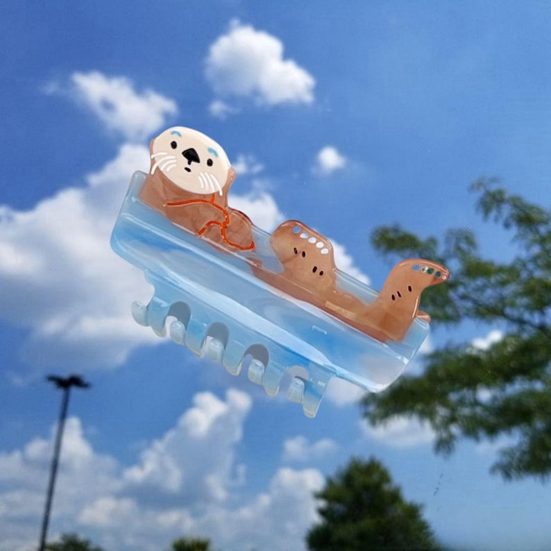 May include: A light blue hair claw clip featuring a brown sea otter design. The clip has a translucent blue base with a smooth finish and a series of teeth along the bottom edge. The otter figure is positioned on top with white and brown details.