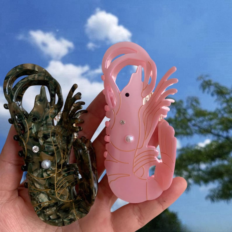 Pode incluir: Dois prendedores de cabelo de pl&aacute;stico em forma de camar&atilde;o. Um &eacute; rosa e o outro &eacute; verde e marrom com detalhes dourados.