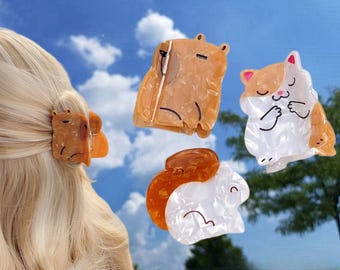 May include: Four colorful hair clips featuring cartoon animals. The clips are made of a translucent plastic with a marbled effect. The animals are a brown capybara, a white cat with orange ears, a white rabbit with brown ears, and a brown squirrel.