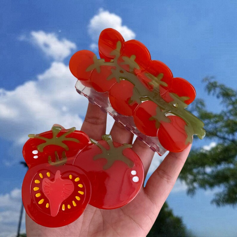 May include: Two red tomato-shaped hair clips. One clip is a single tomato with a slice cut out of it. The other clip is a cluster of tomatoes on a vine.