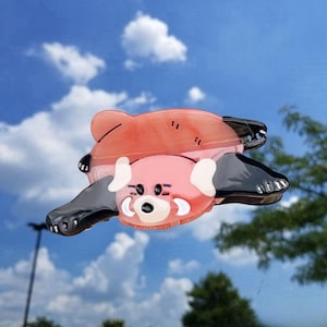 May include: A hair claw clip shaped like a red panda, featuring a translucent reddish-orange body with black limbs and white facial markings. The design includes a dark nose, small eyes, and rounded ears, crafted from a smooth, glossy material.