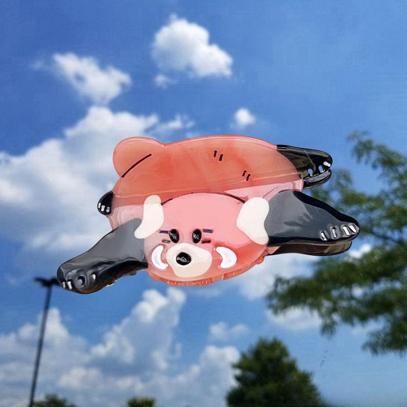 May include: A hair claw clip shaped like a red panda, featuring a translucent reddish-orange body with black limbs and white facial markings. The design includes a dark nose, small eyes, and rounded ears, crafted from a smooth, glossy material.