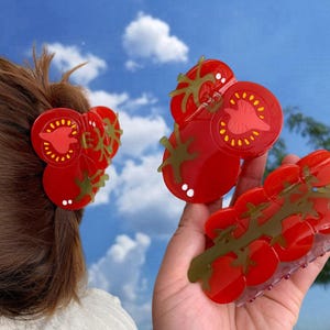 May include: Two red tomato-shaped hair clips. One clip is a single tomato with green leaves. The other clip is a cluster of five tomatoes with green leaves.