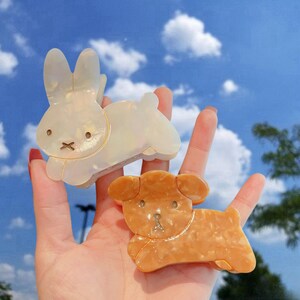 May include: Two hair clips shaped like a bunny and a puppy. The bunny is white with a marbled texture and the puppy is orange with a marbled texture.