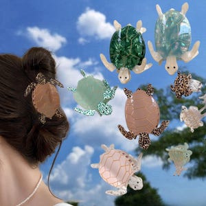 May include: A collection of turtle-shaped hair clips in various colors and patterns. The clips feature a tortoiseshell design, with some having a pearlescent or glittery finish. The background shows a blue sky with clouds and a glimpse of green foliage.