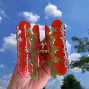 May include: A red and green plastic hair claw with a floral design. The claw is open and ready to hold hair.