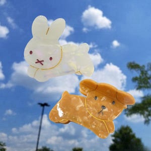 May include: Two plastic hair clips shaped like animals. The first clip is a white rabbit with gold accents. The second clip is a brown dog with gold accents.
