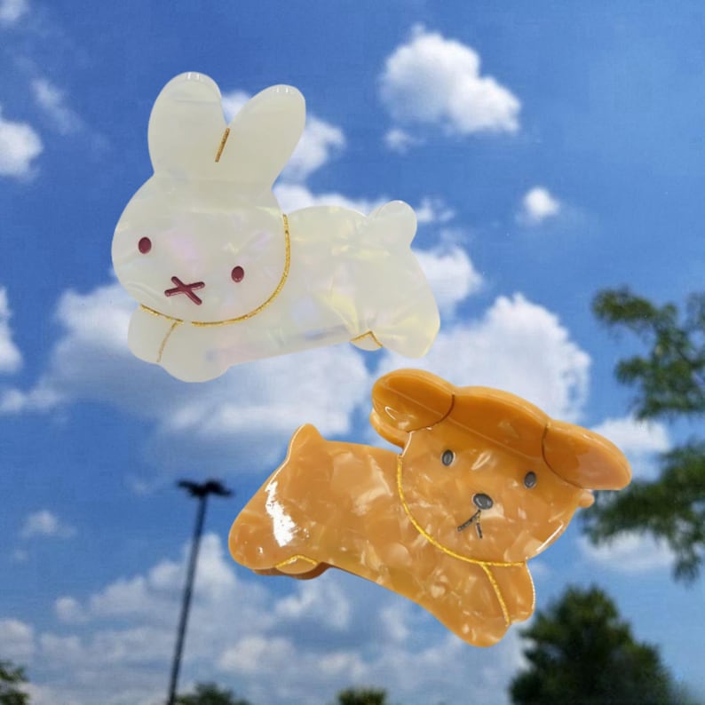 May include: Two plastic hair clips shaped like animals. The first clip is a white rabbit with gold accents. The second clip is a brown dog with gold accents.
