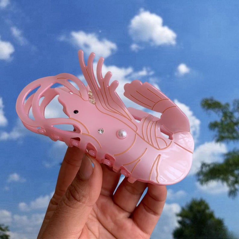 Pode incluir: Presilha de cabelo de pl&aacute;stico rosa em forma de camar&atilde;o com detalhes dourados e enfeites de p&eacute;rolas.