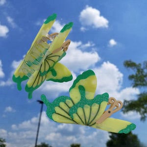 May include: Two yellow butterfly hair clips with green accents. The clips have a textured surface and are designed to hold hair in place.