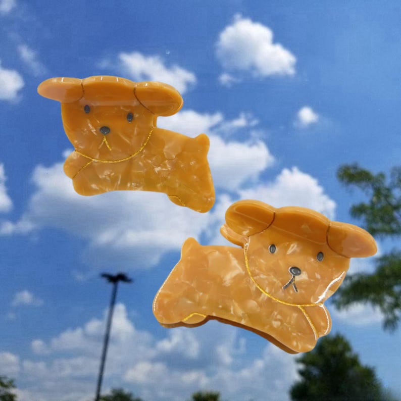 May include: Two yellow plastic hair clips shaped like cartoon dogs. The clips have a marbled effect and gold accents.
