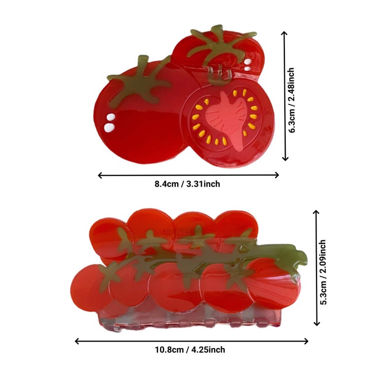 May include: Two red tomato-shaped hair clips. The smaller clip features a single tomato with a green stem and a slice of tomato on top. The larger clip features a cluster of five tomatoes with green stems.