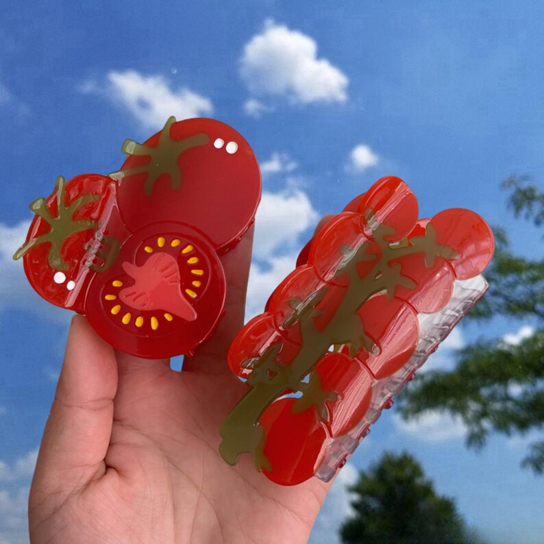 May include: Two red plastic hair clips shaped like tomatoes. One clip is a single tomato, the other is a cluster of tomatoes on a vine.