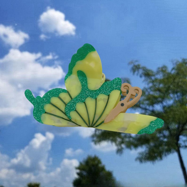 May include: A yellow and green butterfly-shaped hair clip with a glitter finish. The clip has a pair of scissors on the back.