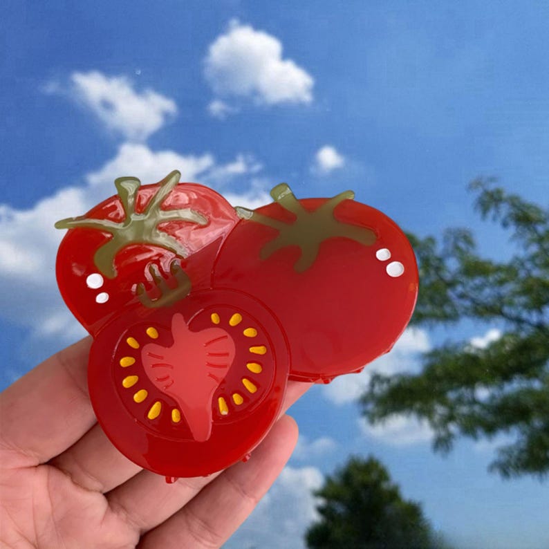May include: Three red tomato-shaped hair clips. One tomato is cut in half and shows the inside of the tomato with yellow seeds. The other two tomatoes are whole.