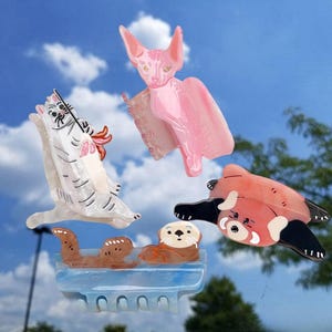 May include: Four hair claw clips featuring animal designs: a white cat with grey stripes, a pink hairless cat, a red panda with black limbs, and two brown otters resting on a light blue base. Each clip has a functional hinge mechanism.