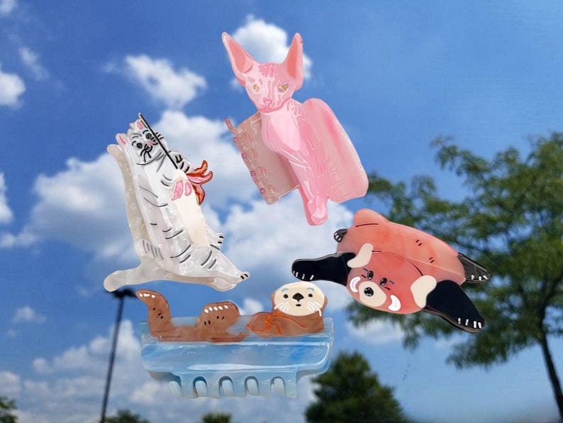 May include: Four hair claw clips featuring animal designs: a white cat with grey stripes, a pink hairless cat, a red panda with black limbs, and two brown otters resting on a light blue base. Each clip has a functional hinge mechanism.