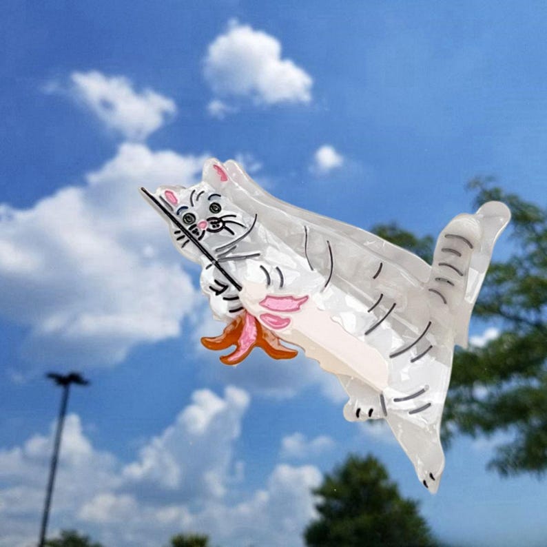 May include: A white cat-shaped hair claw clip with a translucent finish, featuring black line details for the face and body, pink accents on the ears and paws, and a small orange starfish held by the cat, constructed from smooth acrylic material.