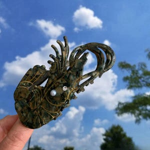 Pode incluir: Uma presilha de cabelo de pl&aacute;stico verde e dourado em forma de lagosta com um enfeite de p&eacute;rola e strass.