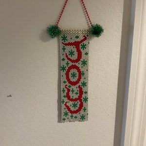 May include: A white beaded banner with the word "JOY" spelled out in red beads. The banner is decorated with green beads and has a red beaded string for hanging.