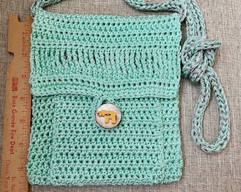 Puede incluir: Un pequeño bolso de hombro de crochet verde claro con un botón de madera con un elefante de dibujos animados. El bolso tiene una larga correa de crochet gris y blanca.