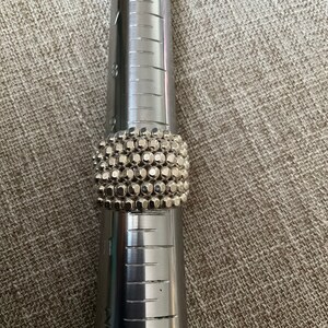 May include: A silver ring with a textured design made of small, round beads. The ring is on a metal ring sizer.