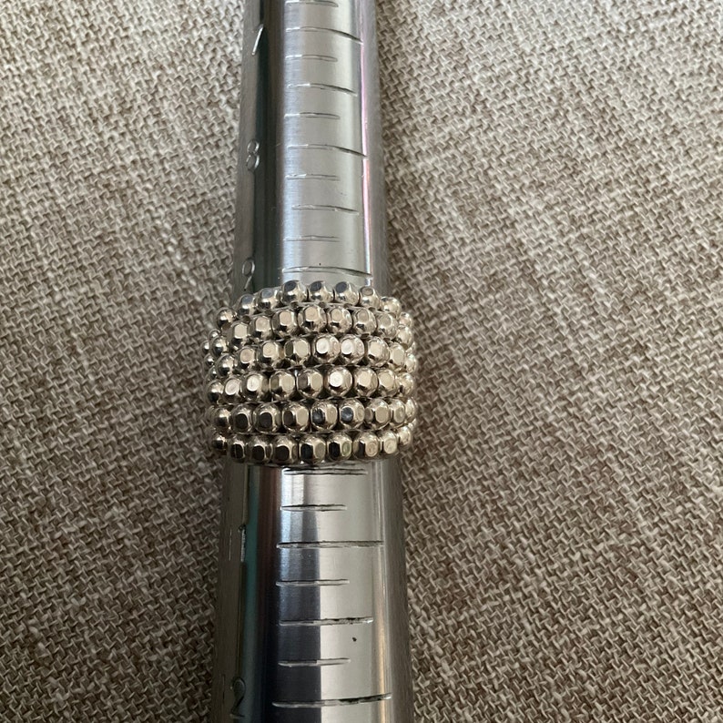 May include: A silver ring with a textured design made of small, round beads. The ring is on a metal ring sizer.