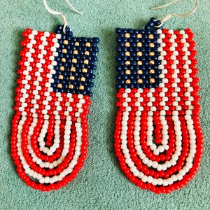 May include: A pair of beaded earrings shaped like the American flag. The earrings are red, white, and blue with gold accents. The earrings are made of small beads and have a silver hook.