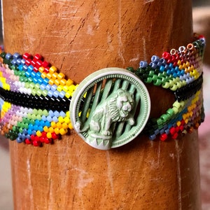 Könnte beinhalten: Ein buntes Perlenarmband mit einem grünen Knopf, der ein weißes Löwenmotiv zeigt.