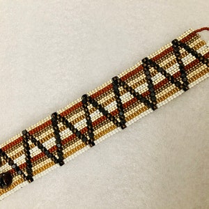May include: A brown, white and black beaded bracelet with a geometric pattern. The bracelet has two brown button closures.