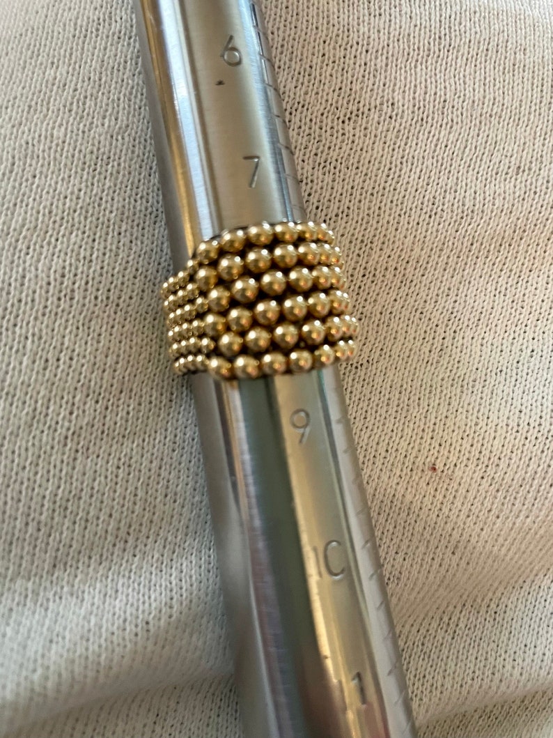 May include: A gold ring made of small beads, measured on a metal ring sizer with markings for sizes 6, 7, 8, 9, 10, and 11.