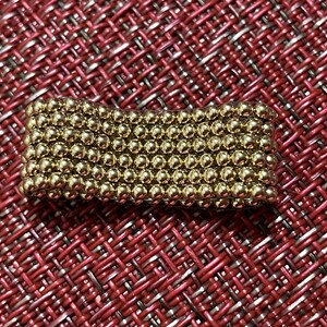 May include: A close-up of a gold-coloured magnetic ball chain. The chain is made up of small, round balls that are connected together. The chain is lying on a red and white patterned surface.
